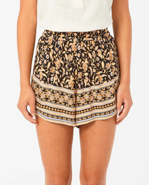 A person wearing Rip Curl Dreamer Short, featuring a black floral pattern with orange and cream details, paired with a white top. The EcoVero/Viscose blend outfit pops against a plain background.