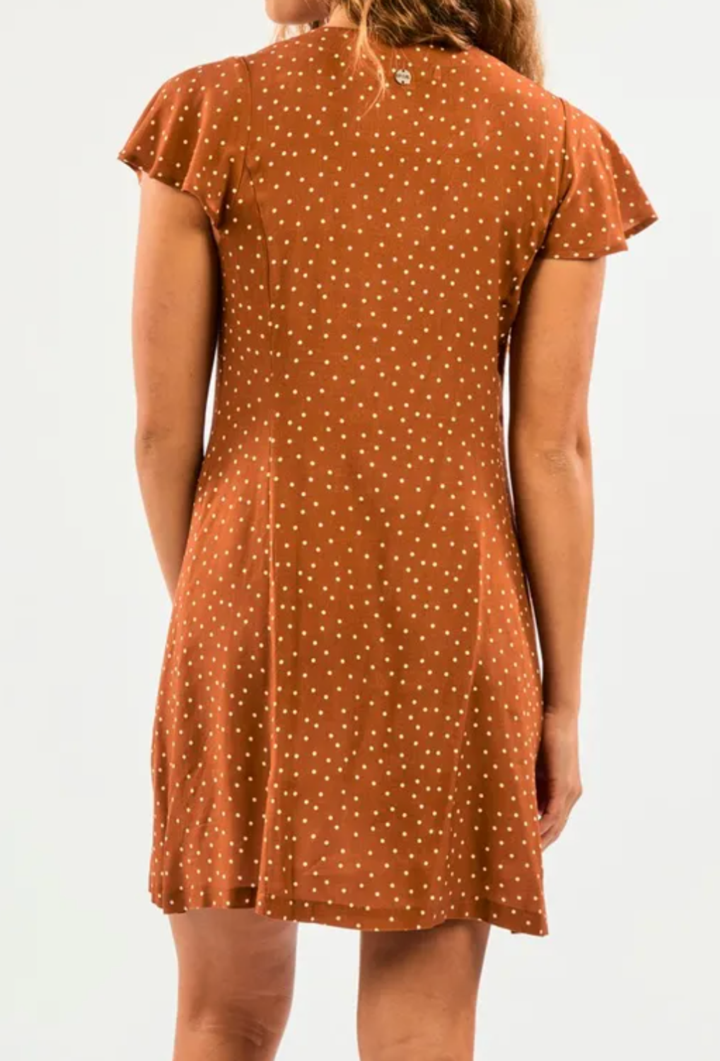 Rear view of a person wearing the Rip Curl Dreamer Spot Button Through Dress, featuring a brown EcoVero/Viscose blend with white polka dots. The knee-length, fitted style complements their wavy hair against a light background.