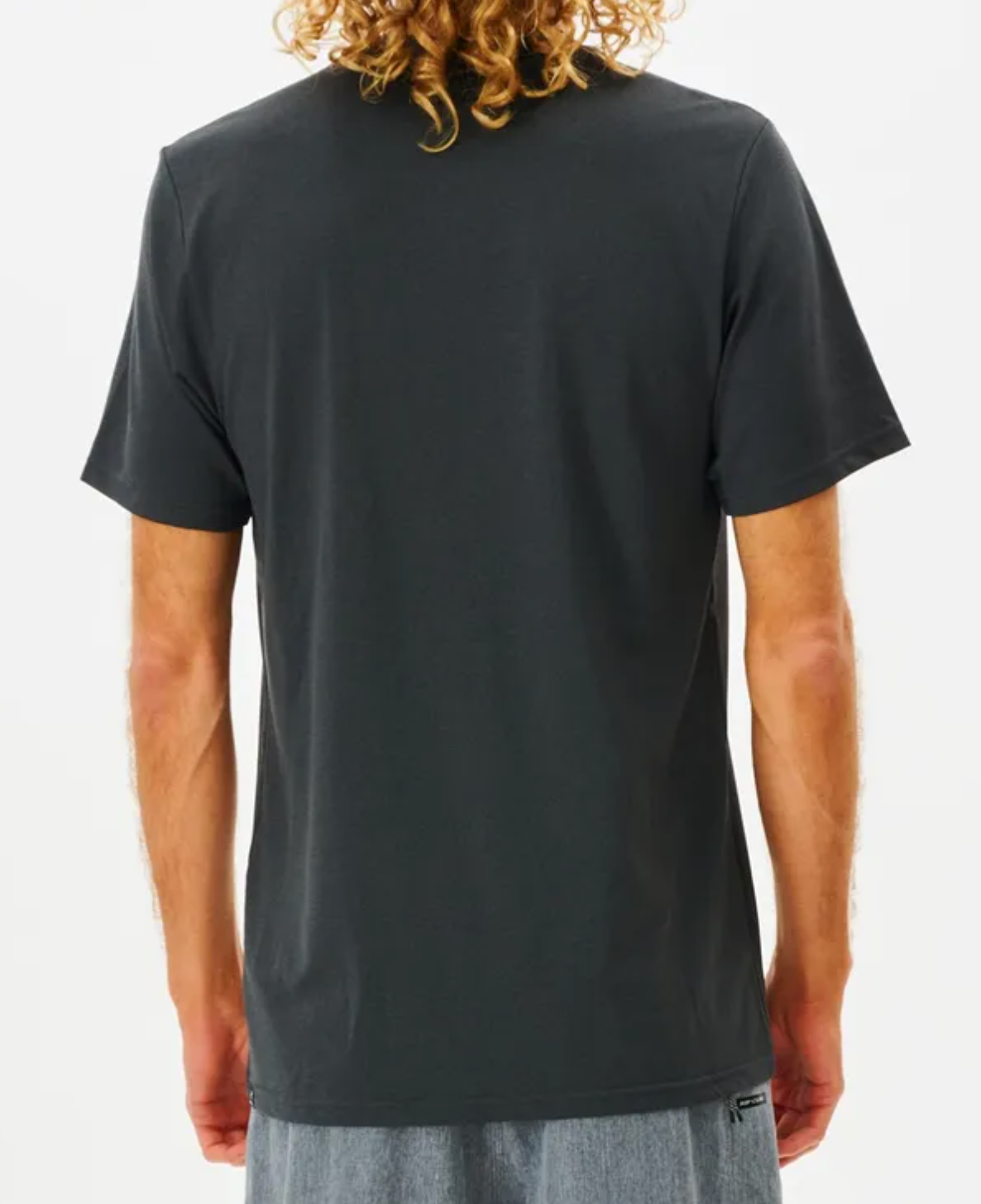 A person with curly hair wears a Rip Curl Search Series Tee, featuring UPF 50+ sun protection in a plain dark design, paired with light-colored pants, viewed from behind against a plain white background.