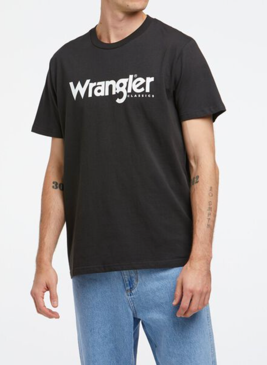Dressed in a Wrangler Classic Logo Tee, 100% cotton and black with "Wrangler" in white across the chest, the person pairs it with light blue jeans on a plain white background.