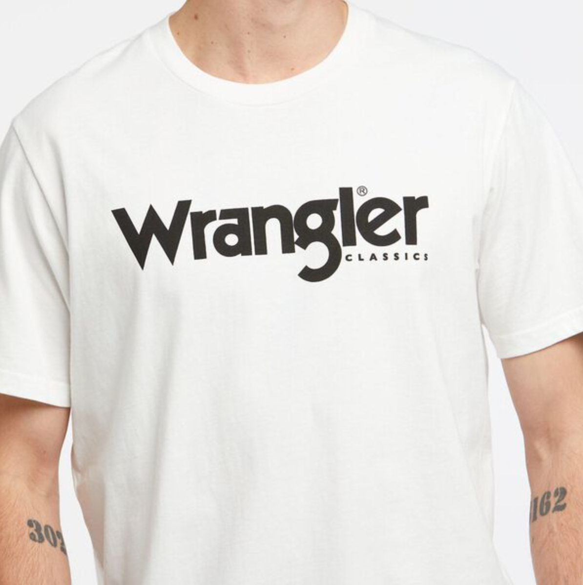 A person wears a Wrangler Classic Logo Tee, featuring "Wrangler" in bold black across the chest. Tattoos of numbers decorate their forearms, with a simple background enhancing the 100% cotton T-shirt's stylish design.