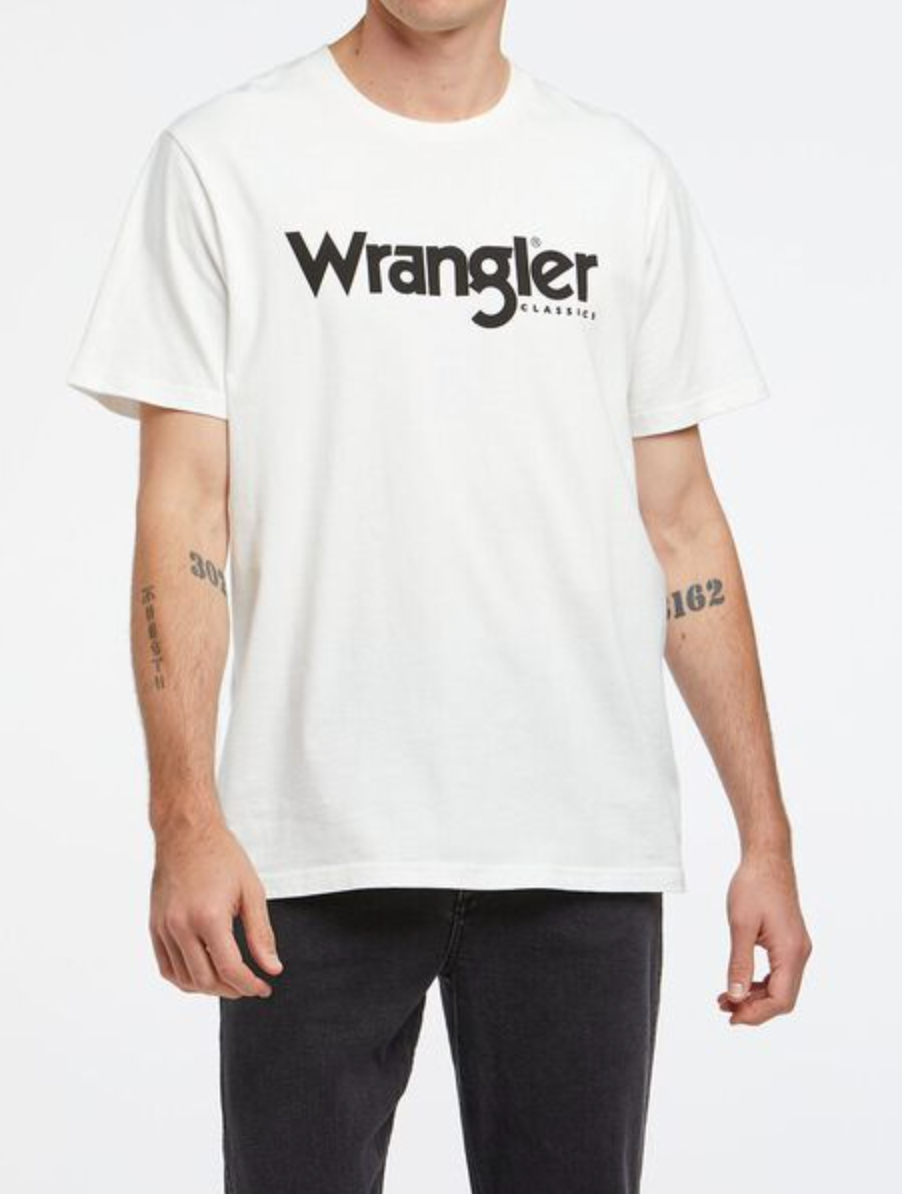 A person sports a Wrangler Classic Logo Tee, with black lettering, 100% cotton. Tattooed forearms complement the look, paired with sleek black jeans against a plain white background.