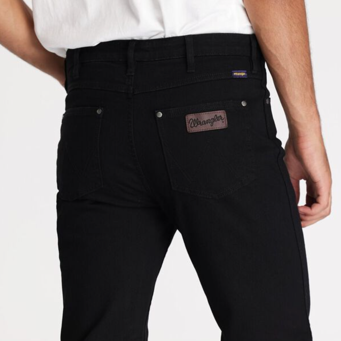 Someone is sporting the Wrangler Classics Straight Jean in black with a white shirt. The focus is on the back of the jeans, showcasing the brown Wrangler logo patch on the right pocket, while part of their right arm is visible.