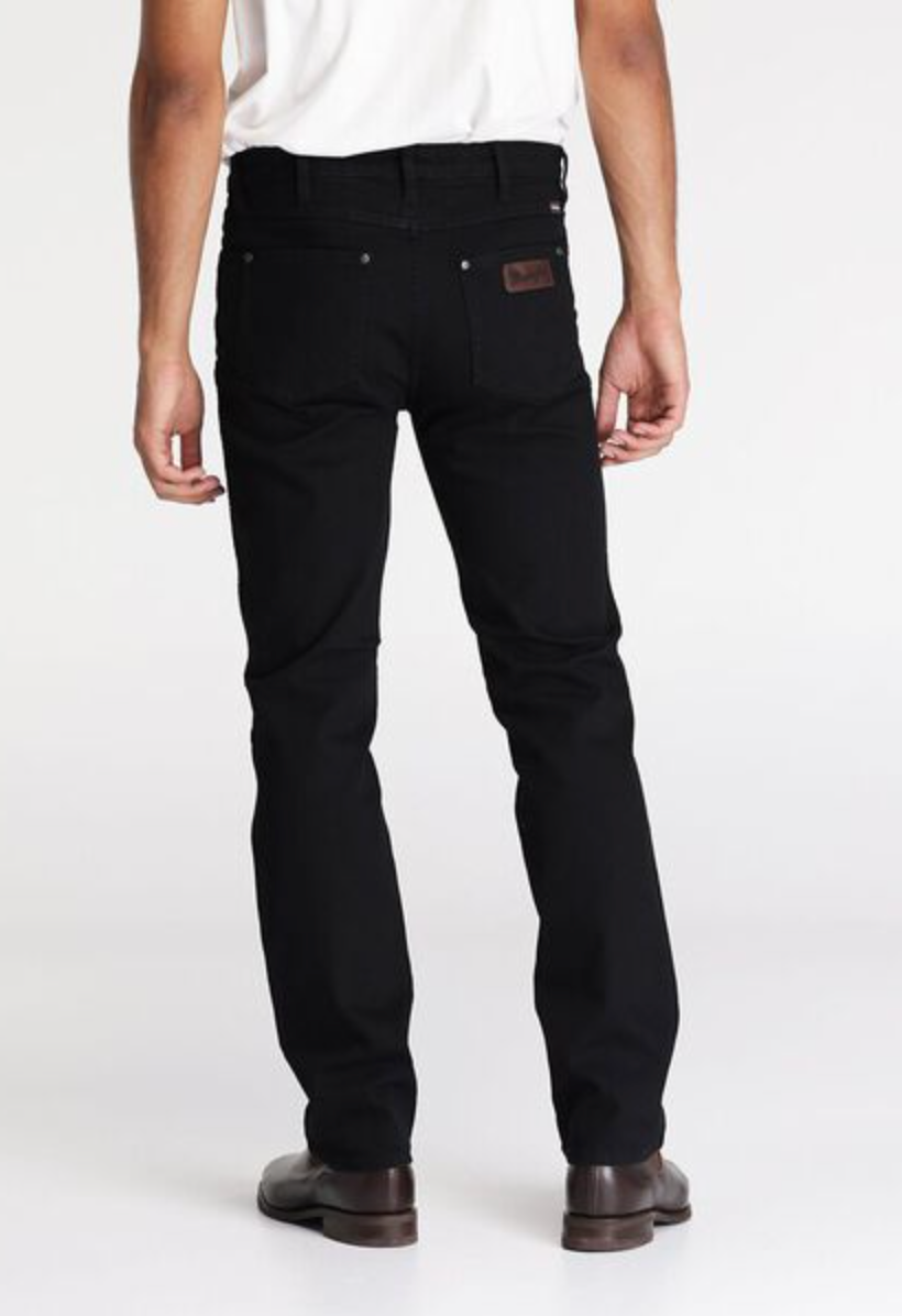 Dressed in Wrangler Classics Straight Jean, a person stands with their back to the camera. They wear a white shirt and brown shoes against a plain white backdrop.