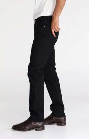 A person is shown from the side wearing Wrangler Classics Straight Jean in black and a white top, with only their lower half visible. They stand on their right leg, left leg slightly bent, wearing brown leather shoes against a plain white background.