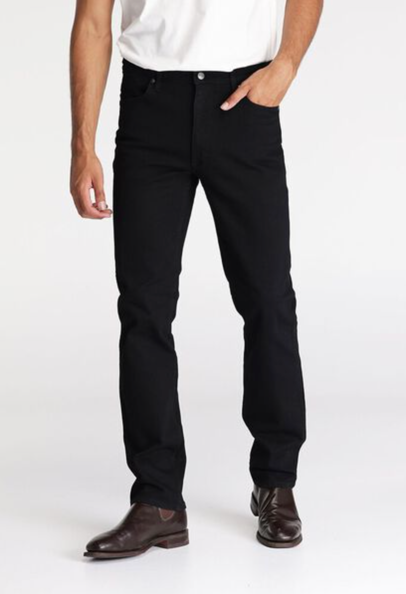A person in a white shirt and black Wrangler Classics Straight Jean stands against a plain backdrop, with one hand casually tucked into the stylish jeans' pocket, complemented by classic brown dress shoes. The focus is on the lower half of the body.