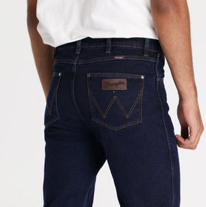 A person is wearing Wrangler Classics Straight Jeans in dark blue, featuring a noticeable brown leather label on the back pocket. They pair these stylish jeans with a white t-shirt, presenting a simple yet fashionable look against the plain white background.