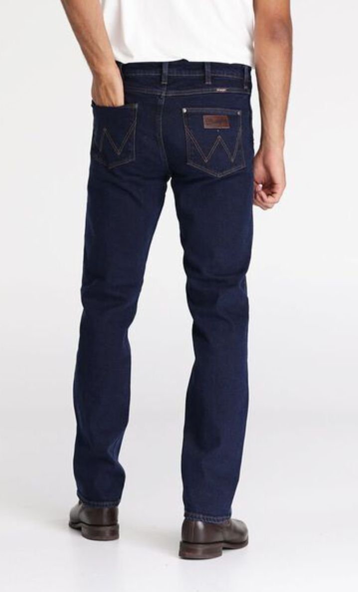 A person wearing a white shirt and dark blue Wrangler Classics Straight Jean stands facing away from the camera. The stylish jeans feature distinctive stitching on the back pockets and a leather patch on the waistband. The person is also sporting dark brown shoes.