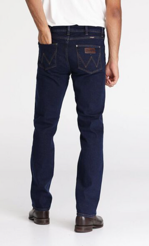 A person wearing a white shirt and dark blue Wrangler Classics Straight Jean stands facing away from the camera. The stylish jeans feature distinctive stitching on the back pockets and a leather patch on the waistband. The person is also sporting dark brown shoes.