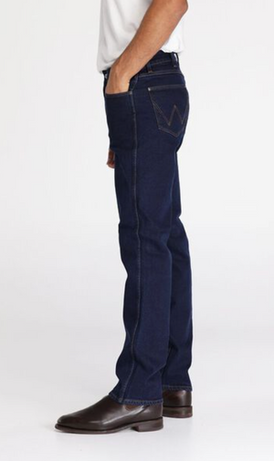 A person is standing sideways with their hand in the pocket of dark blue, comfortable Wrangler Classics Straight Jeans. They are wearing a white shirt and brown leather shoes. The stylish jeans feature contrasting stitching and a "W" design on the back pocket. The background is plain white.