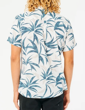 A back view features a person with curly hair wearing the Rip Curl Angourie Short Sleeve Shirt in a blue and white tropical leaf pattern, paired with black pants. The 100% cotton fabric contrasts against the plain white background.
