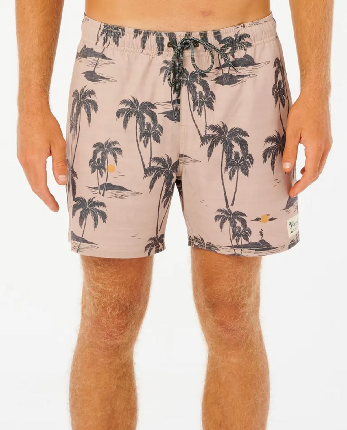 A person in Rip Curl Mod Tropics Volley shorts, light brown with a black palm tree pattern, featuring quick-dry fabric, drawstring waist, and a small logo patch near the hem. They have light skin against a plain white background.