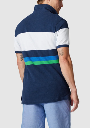 A person wearing the Rodd & Gunn Willow Park Polo, crafted from Australian Cotton with navy and green stripes, stands facing away. Their short hair highlights their arm tattoos, paired with light blue shorts for a relaxed look.
