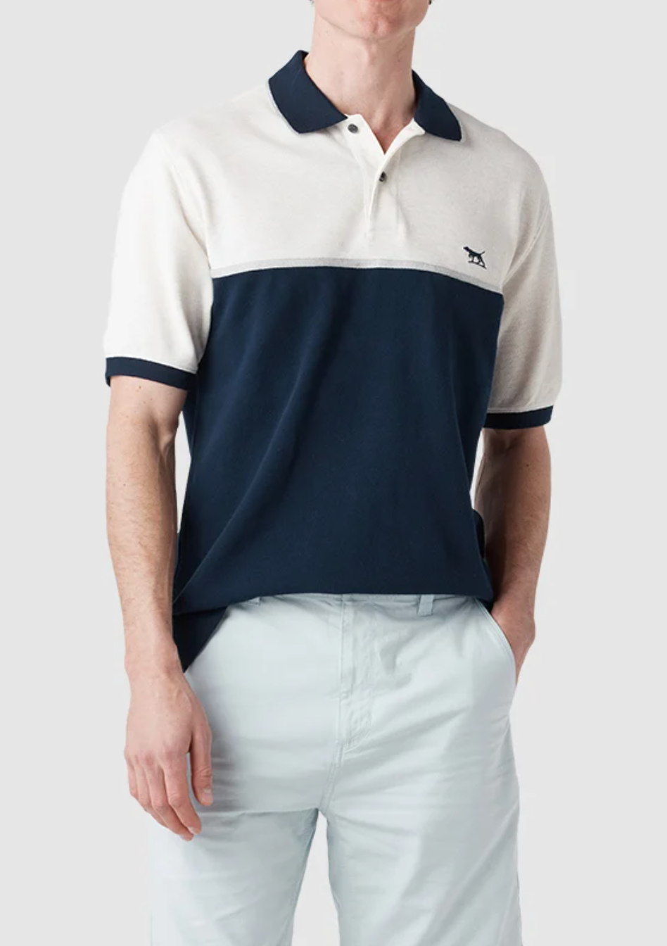 A person is wearing a Rodd & Gunn Castleford Polo, featuring a white top and navy bottom with a chest logo, crafted from premium Australian cotton. They pair it with light-colored pants and have one hand in their pocket.