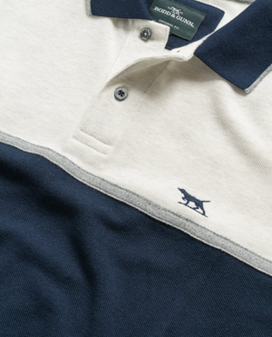 Close-up of the Rodd & Gunn Castleford Polo, a premium two-tone Australian cotton shirt with a beige top and navy bottom. Features include a small embroidered animal logo, "Rodd & Gunn" collar tag, and two visible neckline buttons.