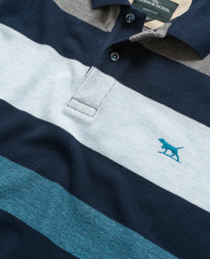 The Rodd & Gunn Lake Hayes Polo features a sports fit with dark blue, white, and light blue stripes. It's crafted from soft Australian cotton and includes an embroidered blue dog logo on the left chest. The polo has a dark blue collar with two matching buttons.