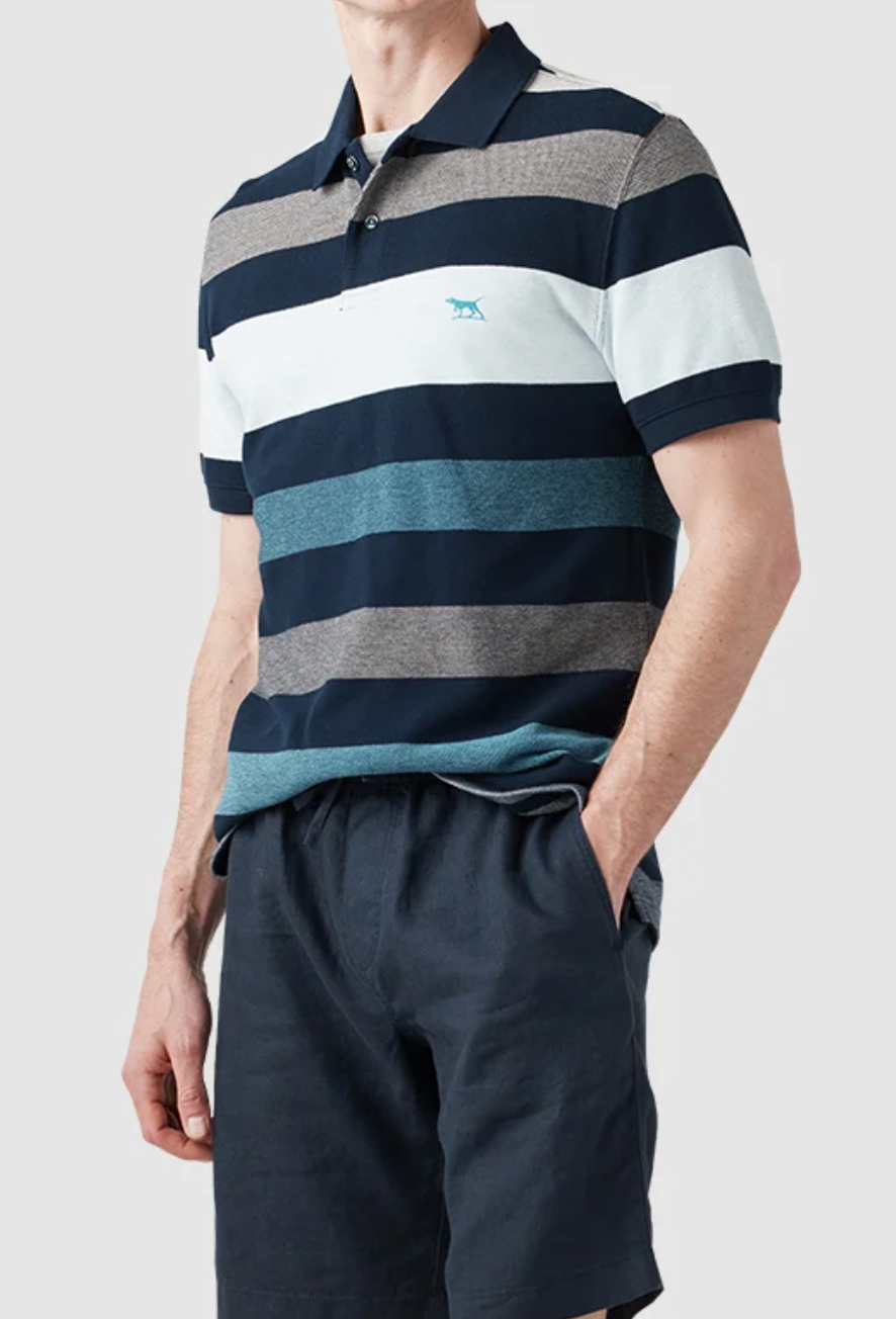 A person wears a Rodd & Gunn Lake Hayes Polo in sports fit with navy, light blue, and gray stripes plus an embroidered design. Paired with dark blue shorts, they stand against a gray backdrop showcasing style crafted from Australian cotton. Hands rest in pockets for a relaxed look.