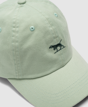 The Rodd & Gunn Gunn Cap in light green is made of 100% Heavy Duty Cotton Twill and features an embroidered pointing dog on the front. It includes a curved brim, visible stitching, and an adjustable strap for a perfect fit, all displayed on a gray surface.