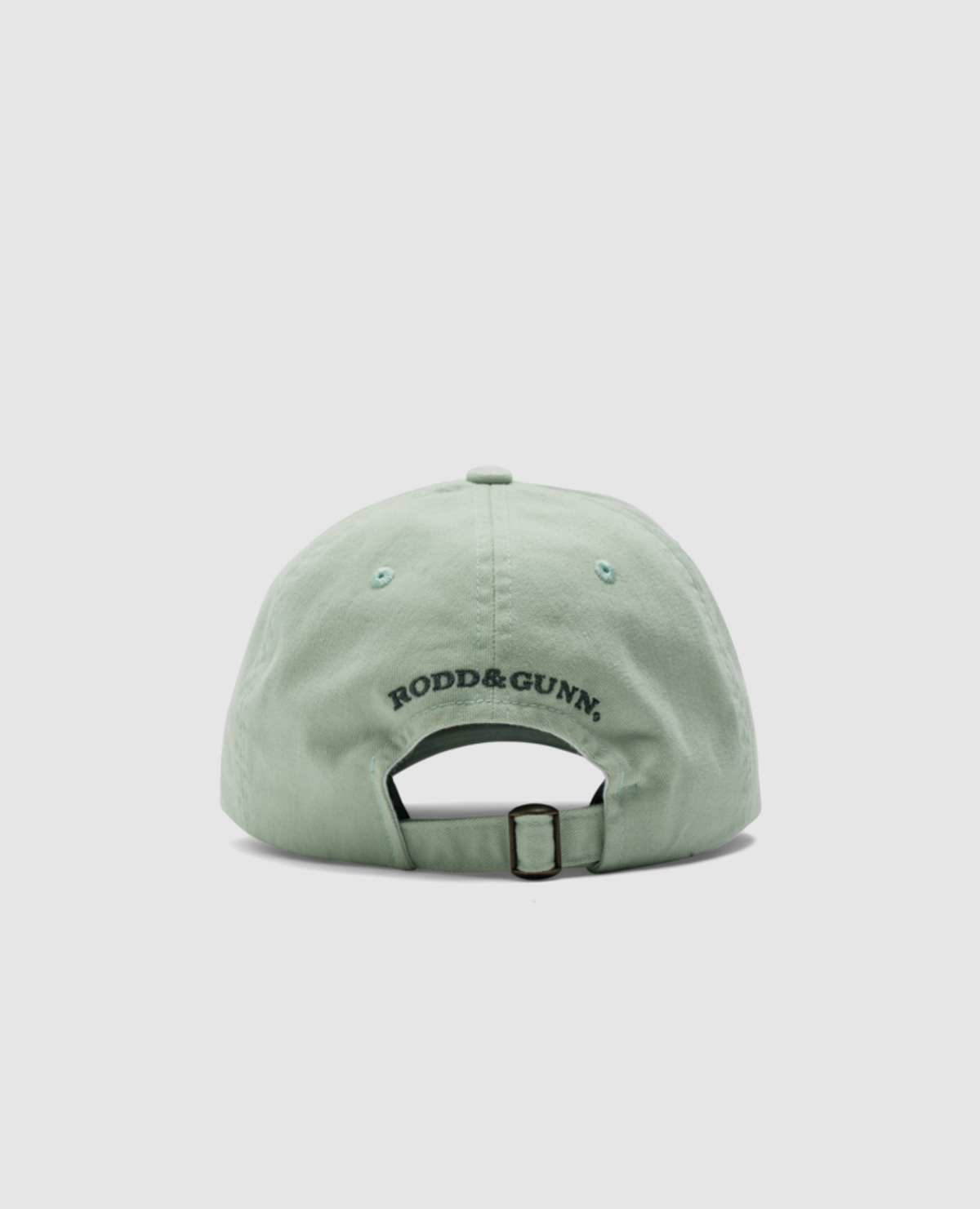 The Rodd & Gunn Gunn Cap is pale green, crafted from 100% heavy-duty cotton twill. "RODD & GUNN" is embroidered in black on the back. It includes an adjustable strap with a metal buckle for a perfect fit.