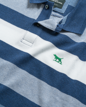 Close-up of a Rodd & Gunn Spencerville Polo, made from Australian cotton, showcasing blue and white stripes with a small green dog logo embroidered on the chest. The polo has a blue collar, with the top button undone, revealing the brand tag inside.