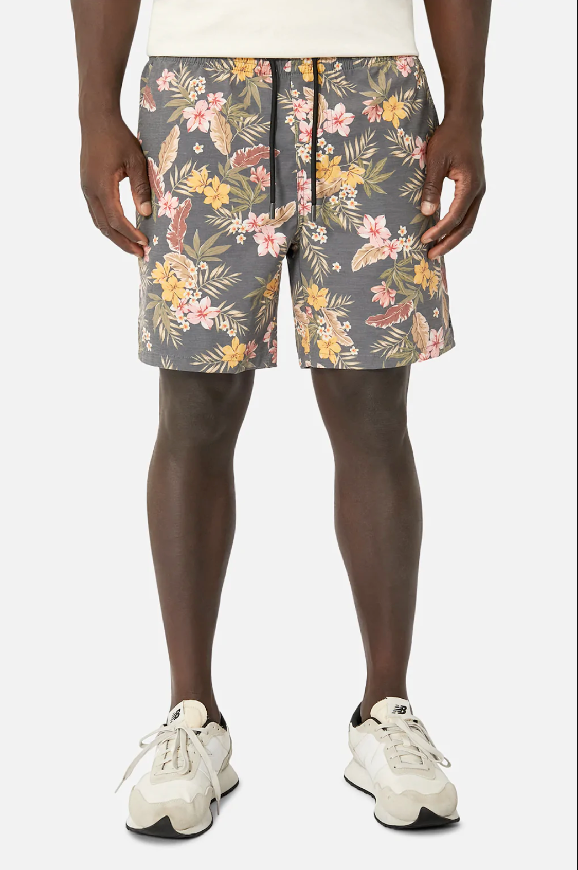 A person wears Industrie The Livadi Bahama floral shorts with pink, yellow, and green tones, featuring an elasticized waist, paired with a crisp white shirt and white sneakers. They stand against a plain white background.