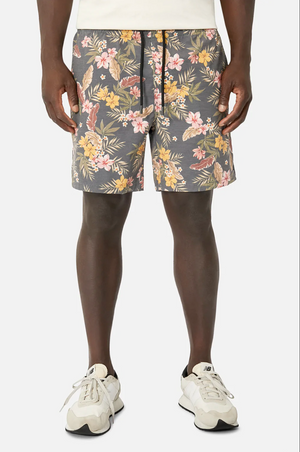 A person wears Industrie The Livadi Bahama floral shorts with pink, yellow, and green tones, featuring an elasticized waist, paired with a crisp white shirt and white sneakers. They stand against a plain white background.
