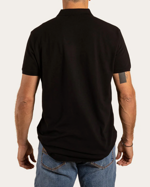 The rear view shows a person in a Ringers Western Essential Polo, black and short-sleeved, made of 100% cotton, paired with blue jeans. A tattoo decorates their left upper arm against a plain, light-colored background.
