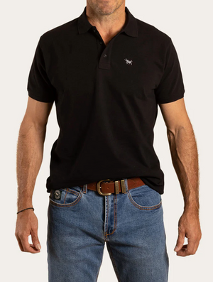 A man wears a Ringers Western Essential Polo in black, crafted from 100% cotton with an embroidered chest logo. He pairs it stylishly with blue jeans and a brown leather belt, his hands resting by his sides against a plain white background.
