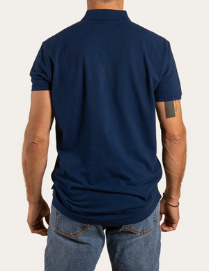 A man, facing away from the camera, wears a navy Ringers Western Essential Polo made of 100% cotton and blue jeans. His arms hang naturally at his sides, revealing a tattoo on his left forearm against the plain white backdrop.