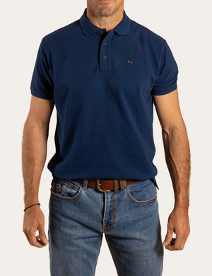 A man stands against a plain background, wearing a Ringers Western Essential Polo in navy blue, made from 100% cotton, paired effortlessly with blue jeans and a brown belt.
