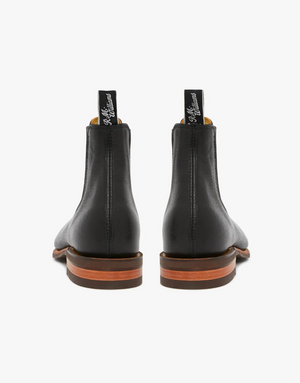 Revised Sentence: Rear view of a pair of RM Williams Comfort Kangaroo Craftsman Boots in black leather with brown soles. The elastic-sided boots feature pull tabs at the back, adorned with white script text. Handcrafted in Australia from premium materials, the image against a white background emphasizes their sleek, polished appearance.