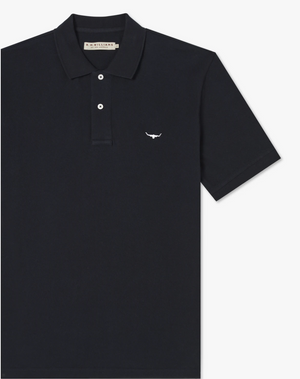 The RM Williams Rod Polo is a black cotton pique polo shirt with a folded collar, featuring a small white longhorn embroidery on the left chest. It has short sleeves, R.M.Williams branded buttons on the three-button placket, and a beige tag with branding information inside the collar.