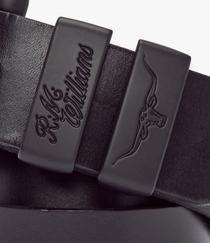 Close-up image of a black RM Williams Drover 1 1/2 Belt featuring a textured, vegetable-tanned leather finish. The belt has two fixed loops: one embossed with the RM Williams logo and the other with a horned bull head design.