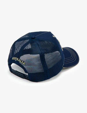 The RM Williams Cremorne Cap is a navy blue trucker hat with a mesh back and an adjustable strap. It features yellow "AUSTRALIA" embroidery on the back and has light-colored stitching along the edge of the brim, adding a touch of style to this classic design.