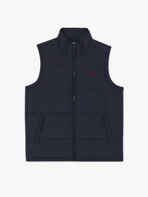 The RM Williams Patterson Creek Vest is a navy blue, sleeveless quilted puffer vest featuring a high collar and a zippered front. It includes two side pockets and displays a small red logo on the left chest area. Crafted from water and wind-resistant Taslan nylon, the material is designed with padding for insulation.