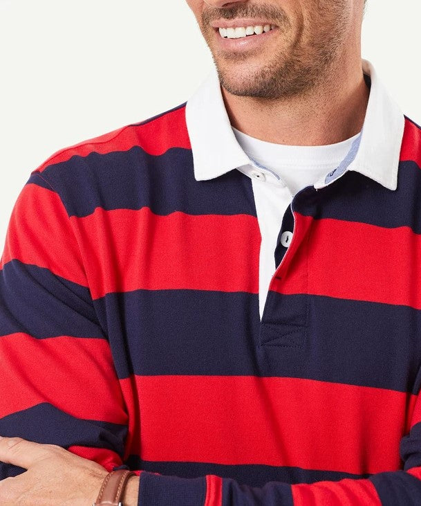 A smiling man with a beard, arms crossed, is wearing a Gazman Block Stripe Rugby jumper in red and navy stripes with a white collar and a white t-shirt underneath. The background is plain and light-colored.