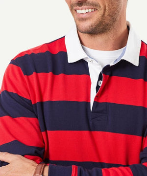 A smiling man with a beard, arms crossed, is wearing a Gazman Block Stripe Rugby jumper in red and navy stripes with a white collar and a white t-shirt underneath. The background is plain and light-colored.