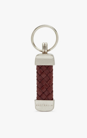 The RM Williams Plaited Key Ring features a silver metal ring and end caps, with a central section of dark brown braided Kangaroo leather. The bottom metal cap is engraved with the word "AUSTRALIA." Accentuated by brass-toned fittings, this keychain boasts a simple and elegant design.