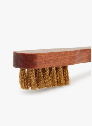 A close-up image of the RM Williams Suede Brass Bristle Brush reveals a wooden handle with dense, short brass bristles. The rectangular handle features a smooth finish, and the bristles appear sturdy for heavy-duty cleaning of suede boots. The background is plain white.
