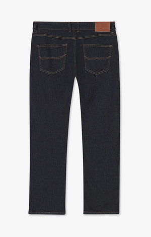 A pair of RM Williams Ramco Jeans in dark blue, premium denim is laid flat, showcasing the backside. The straight-leg jeans feature two back pockets with brown stitching and a leather patch on the waistband, along with belt loops for added functionality.
