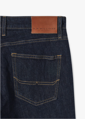 Close-up of the back of a pair of RM Williams Ramco Jeans in dark blue stretch denim, showcasing a single back pocket with yellow stitching and a brown leather patch above it embossed with the text "R.M. WILLIAMS THE BUSH OUTFITTER SINCE 1932 AUSTRALIA." The straight leg jeans are displayed against a white background.