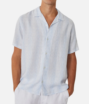 Against a plain white background, a man stands confidently with his hands in pockets, wearing the Industrie The Tropicana Shirt, a light blue and white striped button-down made from 100% linen. Its relaxed fit complements his crisp white pants perfectly.