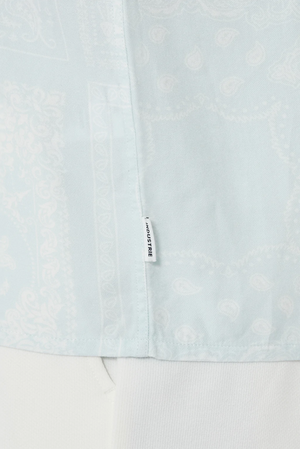 Close-up of a light blue fabric with a subtle paisley and geometric print. A small white tag with black text reads "JACQUEMUS," but the product is actually the Industrie The Stryder Shirt, a relaxed fit button-down. The bottom of the image shows a hint of white fabric.