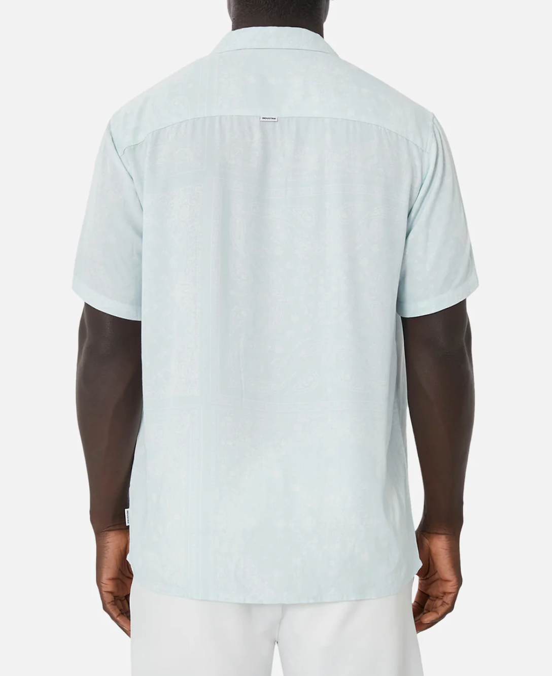 A person is shown from the back wearing the Industrie The Stryder Shirt, a light blue short-sleeve button-down with a subtle paisley pattern. It has a straight hem and relaxed fit, elegantly paired with white pants. The plain white background highlights the outfit effortlessly.
