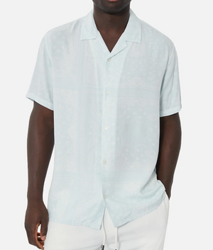 A person in an Industrie The Stryder Shirt, light blue with subtle geometric patterns, stands against a white background. Their left hand is in their pocket and they pair it with relaxed-fit white pants.