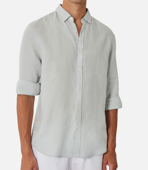 Wearing the relaxed-fit Industrie The Tennyson Linen Shirt made of 100% linen in light gray, their torso is styled with sleeves rolled casually and untucked over crisp white pants, set against a white backdrop, showing only the torso in the frame.