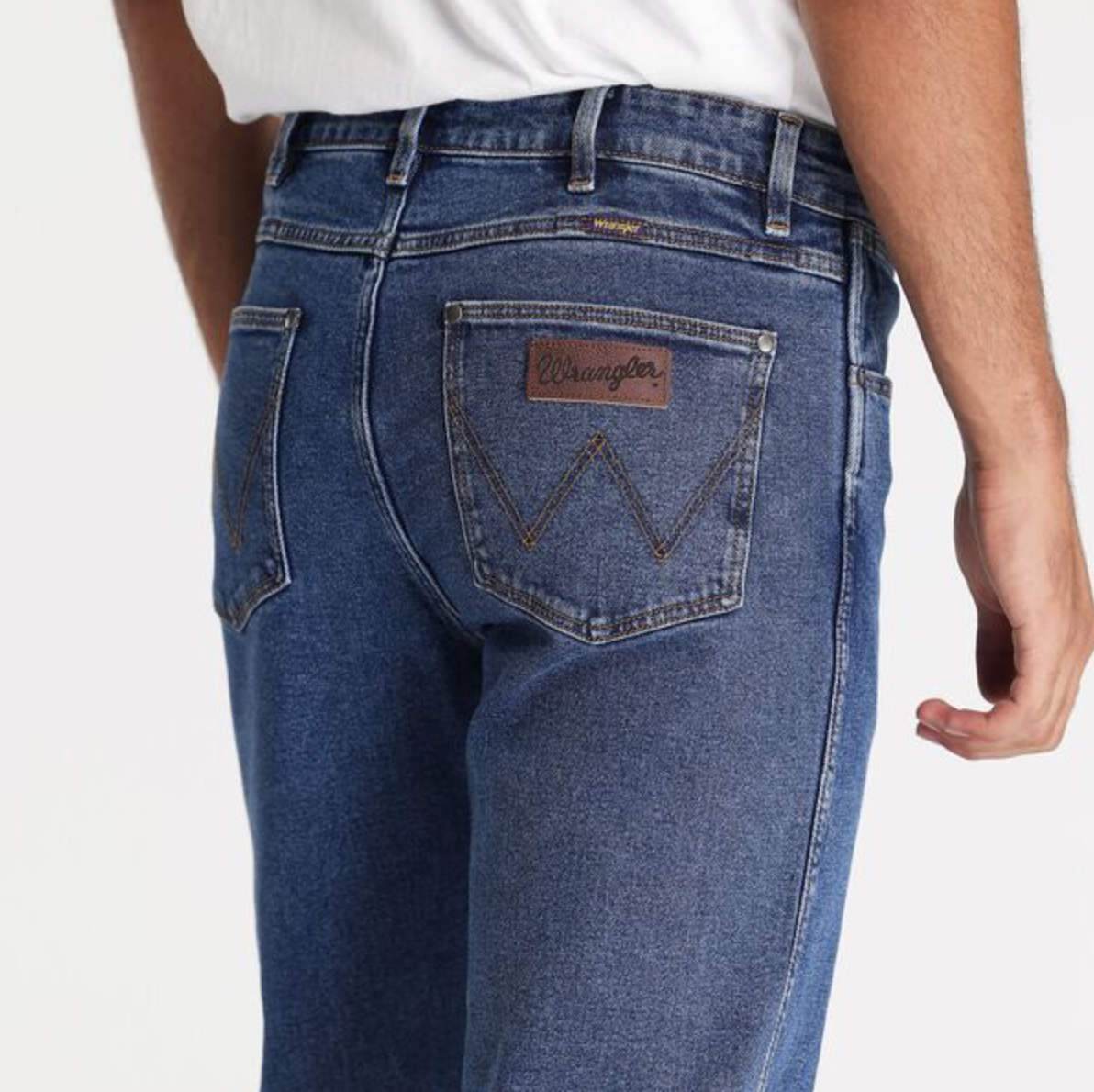 A person is wearing Wrangler Classics Straight Jeans in blue, featuring a mid-rise, straight-leg fit with slight fading around the seams and a brown leather patch with the Wrangler logo on one back pocket, paired with a white shirt. The perspective shows them from the back, below the waist.