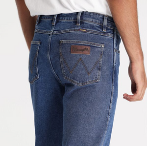A person is wearing Wrangler Classics Straight Jeans in blue, featuring a mid-rise, straight-leg fit with slight fading around the seams and a brown leather patch with the Wrangler logo on one back pocket, paired with a white shirt. The perspective shows them from the back, below the waist.