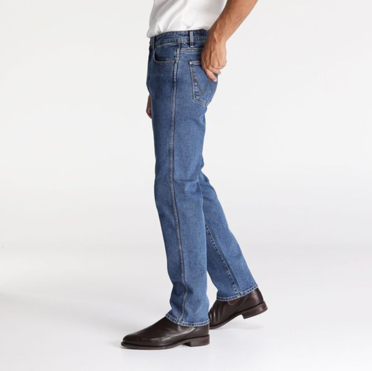 A person wearing Wrangler Classics Straight Jeans, a white shirt, and brown leather shoes stands against a plain white background. Only the lower half of the body is visible, with one hand in a pants pocket.