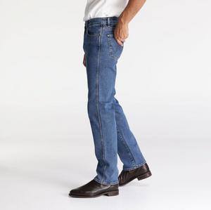 A person wearing Wrangler Classics Straight Jeans, a white shirt, and brown leather shoes stands against a plain white background. Only the lower half of the body is visible, with one hand in a pants pocket.